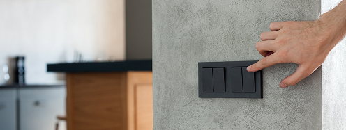 Main appuyant sur un interrupteur noir sur un mur gris, cuisine moderne en arrière-plan. Installation électrique Degee.